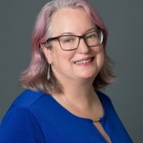 Dr. B with pink hair, wearing a blue shirt and glasses, smiling.